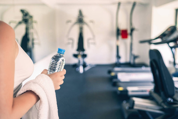 Cropped. Sporty girl with towel on shoulders and bottle still water in the fitness gym opposite the sport equipment and  jogging simulators. Resting time. Health concept. - Foto, Imagen