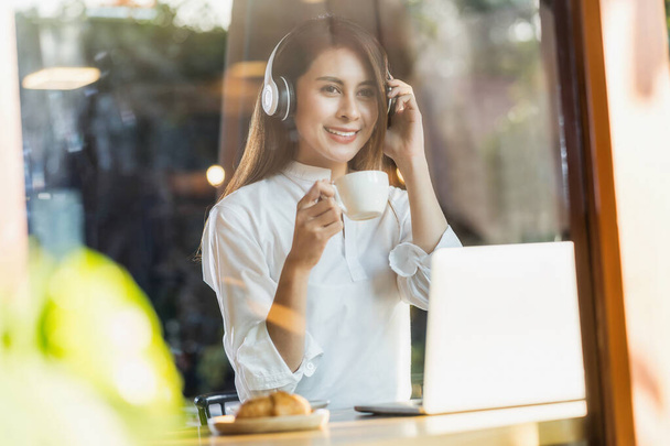 Bir kahve dükkanında elinde bir fincan kahve tutan ve teknoloji laptopunu müzik dinlemek için kullanan genç Asyalı kadın, serbest yazar ve girişimci internete bağlanarak çalışıyor.. - Fotoğraf, Görsel