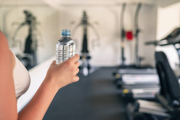 Cropped. Girl hand with bottle still water in the fitness gym opposite the sport equipment and  jogging simulators. Resting time. Health concept. - Photo, image