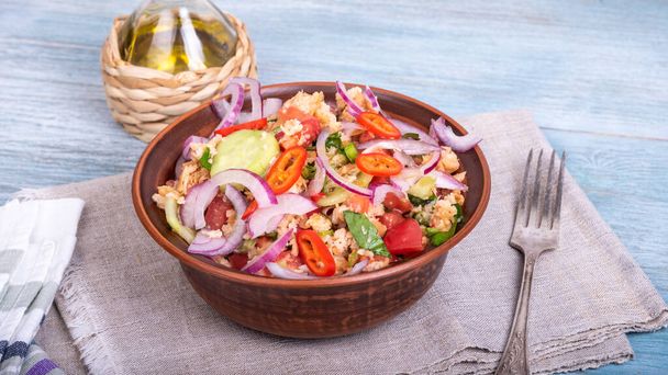 Olasz toszkán vegetáriánus panzanella saláta, vegyes kenyér-, paradicsom- és zöldségsaláta agyagtálban, vászonszalvétán és egy kancsó olívaolaj egy fából készült asztalon. - Fotó, kép