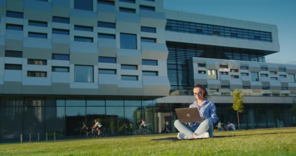 A businesswoman is sitting on the lawn and working on a laptop. Business center in the background. The sun is shining. 4K - Footage, Video