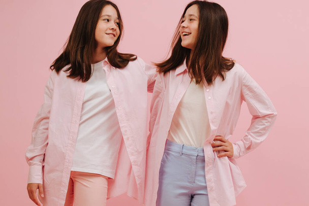 Beautiful little twin sisters happily looking at each other. Over pink background, studio shot. They wear pink and blue pants, similar clothes otherwise. - Photo, image