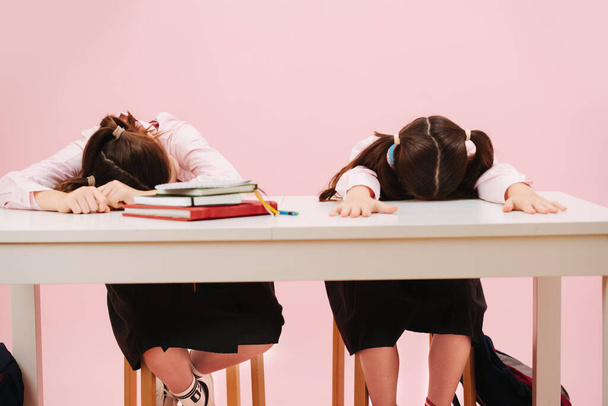 Beautiful little twin schoolgirls sitting behind schooldesk. Frontal view. Over pink background, studio shot. They are bored, sleeping face down. - Photo, image