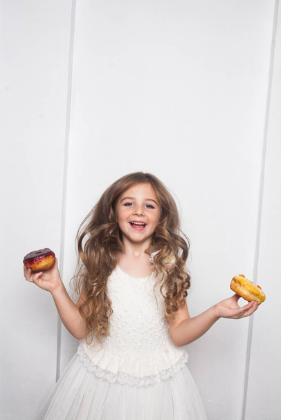 Klein vrolijk schattig kleuter meisje eet kleurrijke donuts op witte achtergrond geïsoleerd. Emotioneel kind heeft plezier met snoep. Lekker zoet eten voor kinderen. Gratis kopieerruimte voor tekst mockup  - Foto, afbeelding