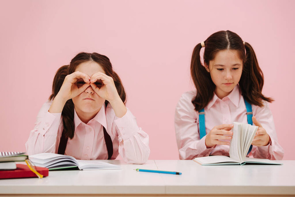 Beautiful little twin schoolgirls sitting behind schooldesk. Frontal view. Over pink background, studio shot. One is bored, making hand binoculars. Second one is flipping pages in textbook. - Foto, afbeelding