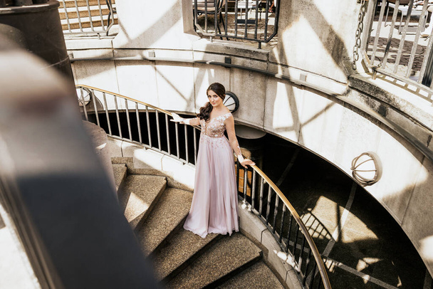 Beautiful attractive woman stand on stairs in pink dress and look in camera - Photo, Image