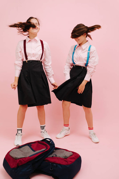 Hermosas colegialas gemelas con gemelos agitando cabezas de lado a lado. Bajo ángulo. Sobre fondo rosa, plano de estudio. Mochilas en el suelo - Foto, imagen