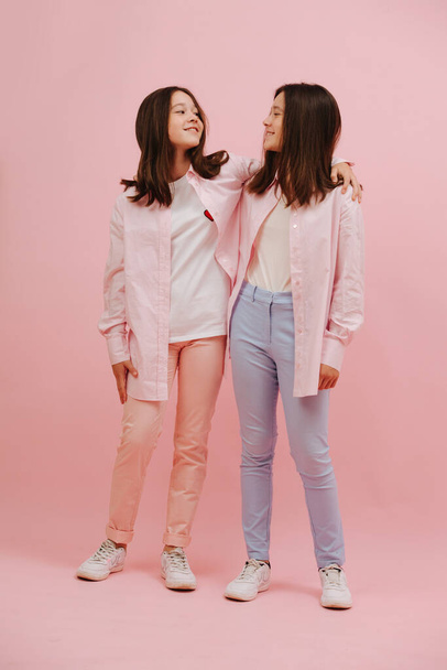 Beautiful little twin sisters happily looking at each other. Over pink background, studio shot. They wear pink and blue pants, similar clothes otherwise. - Fotoğraf, Görsel