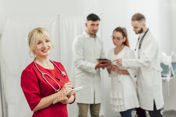 Ofisinde not defteri olan kırmızı üniformalı genç bir doktor. Kameraya bakan kadın. Arka planda, üç doktordan oluşan bir ekip konuşuyor. - Fotoğraf, Görsel