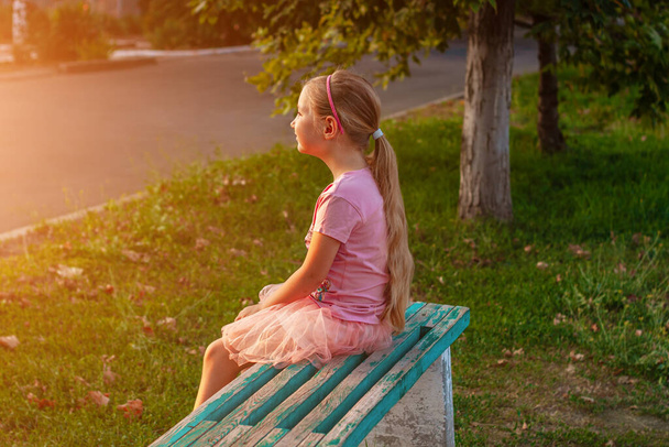 Cheerful little girl with long blonde hair in pink tulle skirt sitting alone on a bench on empty street on sunset. Happy dreamy cute child on nature background. Kids dress fashion. Greeting card. - Photo, Image