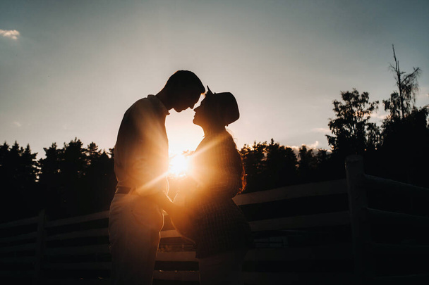 a pregnant girl in a hat and her husband in white clothes stand next to a horse corral at sunset.a stylish couple is waiting for a child in nature. - Fotografie, Obrázek