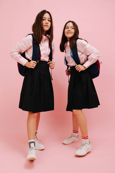 Beautiful little twin schoolgirls happily posing with their backpacks. Low angle. Over pink background, studio shot. Holding straps. One of them looking at camera. - Φωτογραφία, εικόνα