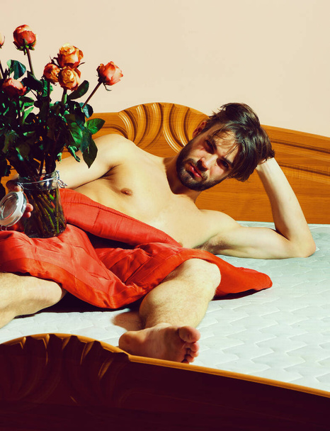 γενειοφόρος άντρας με τριαντάφυλλα στο κρεβάτι κάτω από κόκκινη κουβέρτα κρατώντας διαφανές βάζο - Φωτογραφία, εικόνα