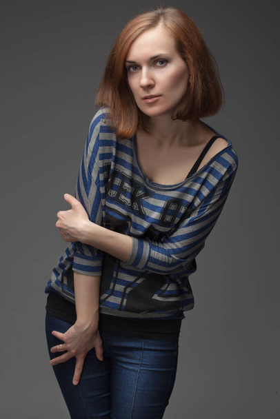 the beautiful girl poses in studio, looks in the camera. red hair, blue jeans, a gray background, an undershirt in lines - Foto, immagini