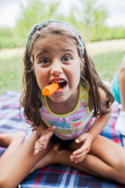 Vicces arckép egy kislányról, aki répát eszik pikniken a parkban a nyári napon. - Fotó, kép
