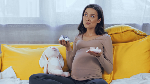 Embarazada mujer holding bebé botines cerca suave juguete en sofá  - Imágenes, Vídeo