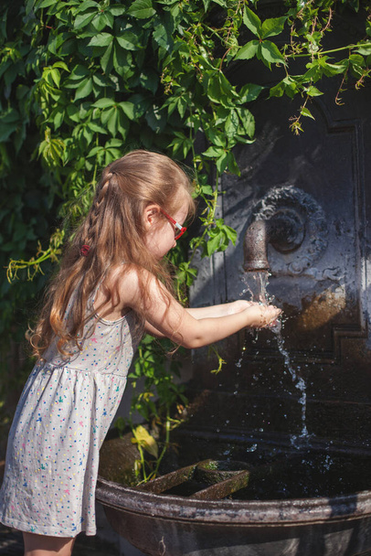 young girl in a point is drinking water and washes her hands in the water in the fresh air. Hungary, Budapest. crane spring in the historical territory of the castle - Fotó, kép