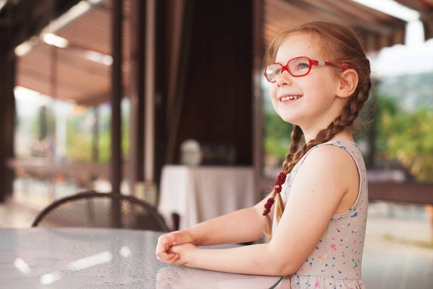 young girl with glasses and dress is sitting at a table in a cafe in the open air - Фото, зображення