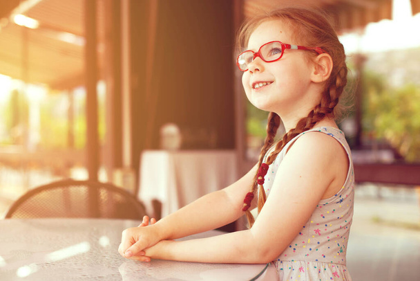 young girl with glasses and dress is sitting at a table in a cafe in the open air - Photo, image
