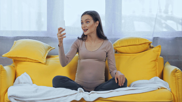 Kobieta w ciąży mając rozmowy wideo na smartfonie na kanapie  - Materiał filmowy, wideo