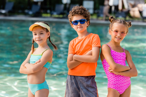 chłopiec w okularach przeciwsłonecznych i dziewczęta w kostiumach kąpielowych pozujących ze skrzyżowanymi ramionami przy basenie - Zdjęcie, obraz