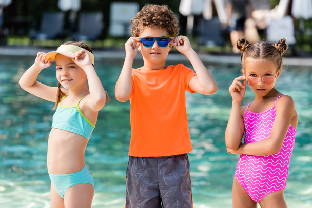 curly boy in t-shirt and girls in swimsuits touching sunglasses near pool - Zdjęcie, obraz