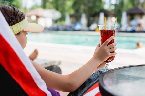 selektywne skupienie dziewczyny w czapce z daszkiem przeciwsłonecznym trzymającej kieliszek koktajlowy podczas odpoczynku na leżaku - Zdjęcie, obraz