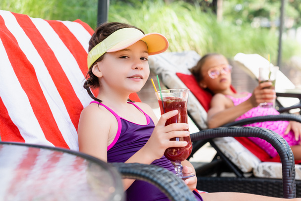 selective focus of girl in sun visor cap sitting in sunbed near friend and holding glass of fresh cocktail - Photo, Image