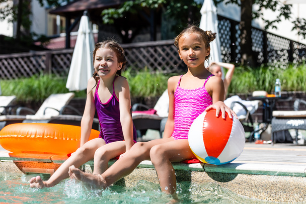 dziewczyny w strojach kąpielowych siedzące w pobliżu basenu z nadmuchiwaną piłką i pierścieniem pływackim podczas wykonywania rozprysków wody z nogami - Zdjęcie, obraz