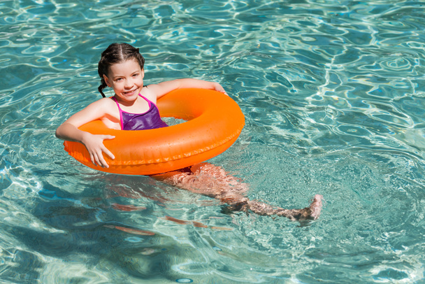 joyful girl looking at camera while floating in pool on swim ring - Photo, Image