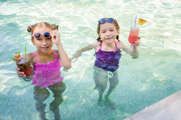 dívky v plavkách s ovocnými koktejly při pohledu na kameru při stání v bazénu - Fotografie, Obrázek