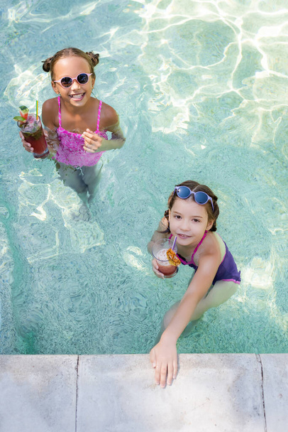high angle view of girls looking at camera while holding glasses with fruit cocktails in pool - Foto, Imagen