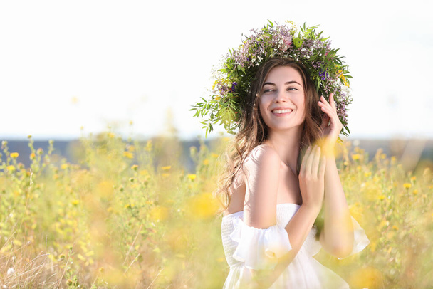 Young woman wearing wreath made of beautiful flowers in field on sunny day - Foto, afbeelding