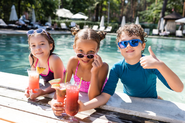 chico en gafas de sol mostrando el pulgar hacia arriba cerca de las niñas con cócteles de frutas frescas en la piscina - Foto, Imagen