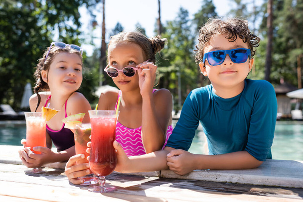dziewczyna w stroju kąpielowym dotykając okulary przeciwsłoneczne w pobliżu przyjaciół z orzeźwiające koktajle na basenie - Zdjęcie, obraz