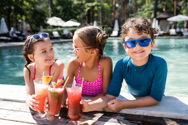 joyful kids in swimwear clinking fresh fruit cocktails near pool - Foto, imagen
