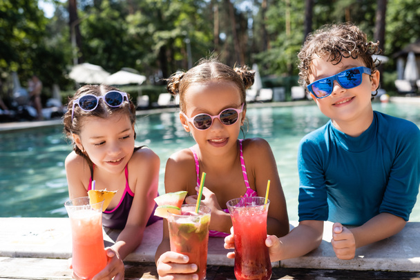 amis joyeux en lunettes de soleil et maillots de bain tenant des cocktails de fruits frais près de la piscine - Photo, image