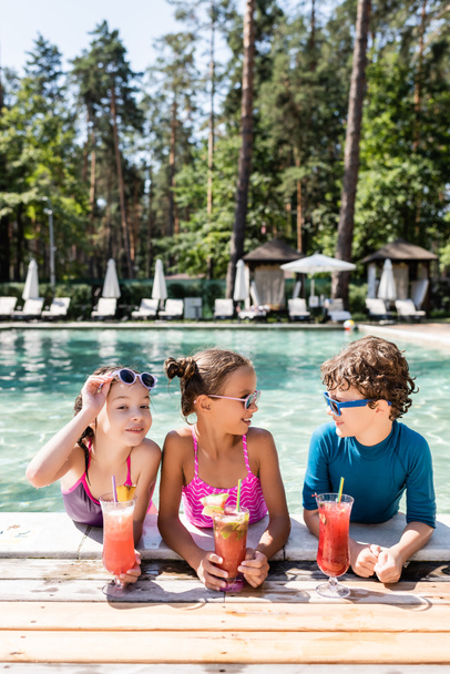 fröhliches Mädchen, das Sonnenbrille anfasst, während es in die Kamera schaut und Freunde mit frischen Fruchtcocktails am Pool anschaut - Foto, Bild