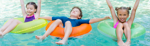 concepto panorámico de las niñas con las manos en el aire y el niño con los ojos cerrados nadando en la piscina en anillos inflables - Foto, Imagen