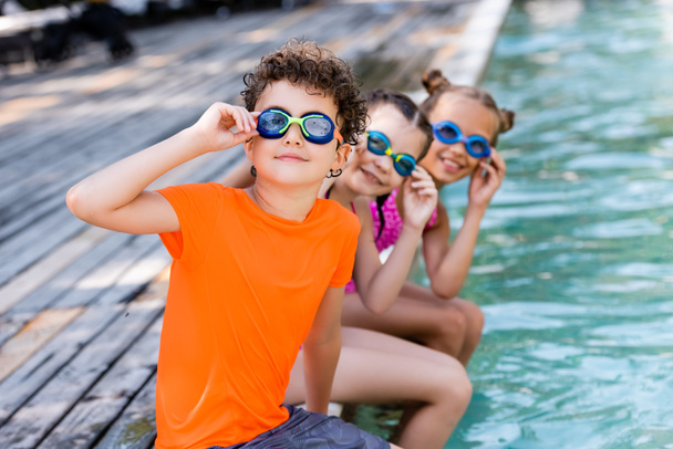 selektivní zaměření přátel dotýkajících se plaveckých brýlí při sezení u bazénu a při pohledu do kamery - Fotografie, Obrázek