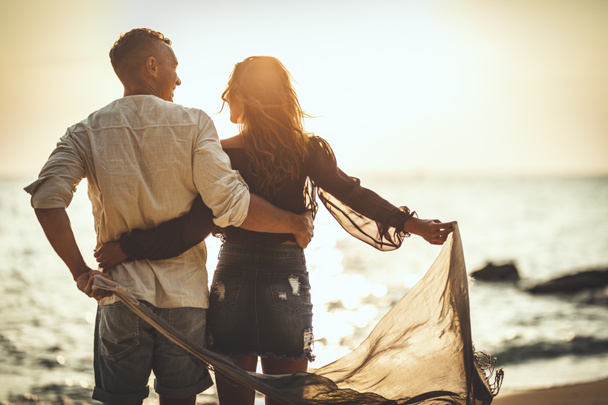 愛するカップルは、夕日の空の砂浜を楽しんで歩いています。彼らはお互いを見ている. - 写真・画像