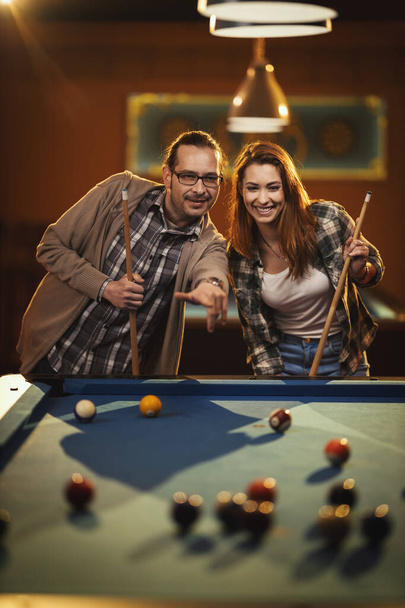 Das junge lächelnde fröhliche Paar spielt nach Feierabend Billard in der Bar. Sie engagieren sich in der Freizeitgestaltung. - Foto, Bild