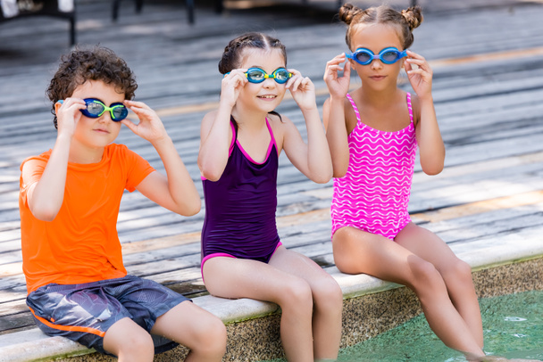 chicas en trajes de baño y niño en camiseta tocando gafas de baño mientras se sienta en la terraza de la piscina - Foto, Imagen