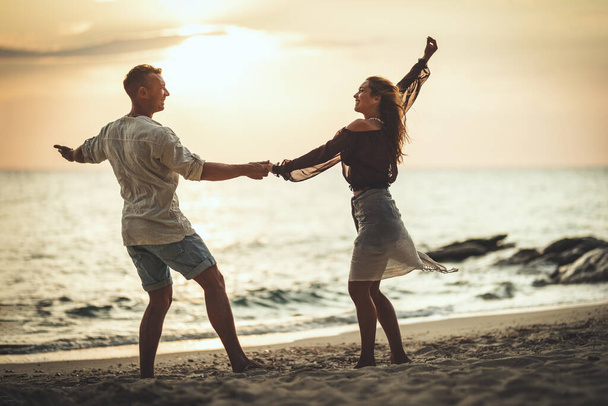 Любляча пара розважається і танцює на порожньому піщаному морському пляжі на заході сонця. Вони дивляться один на одного і щасливо посміхаються
. - Фото, зображення