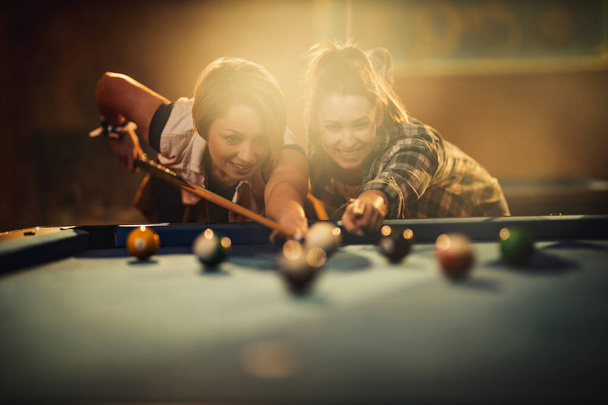 Νεαρή χαμογελαστή χαρούμενα δύο νεαρές γυναίκες παίζουν μπιλιάρδο στο μπαρ μετά τη δουλειά. Συμμετέχουν σε ψυχαγωγικές δραστηριότητες. - Φωτογραφία, εικόνα