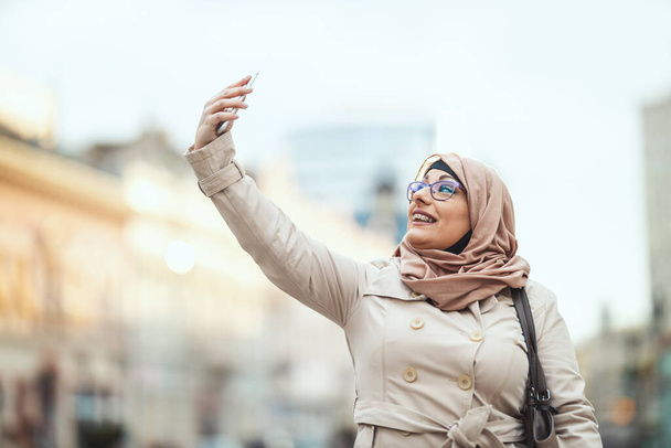 Femme musulmane d'âge moyen portant le hijab avec un visage heureux debout dans l'environnement urbain, faire du selfie par son smartphone. - Photo, image