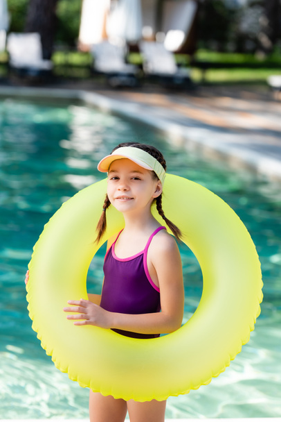 child in sun visor cap and swimsuit posing with inflatable ring near pool - Photo, Image