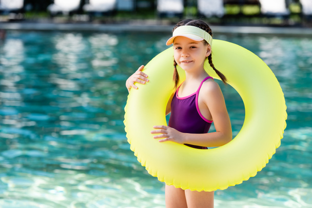 girl in swimsuit and sun visor cap holding inflatable ring while looking at camera near pool - Photo, Image