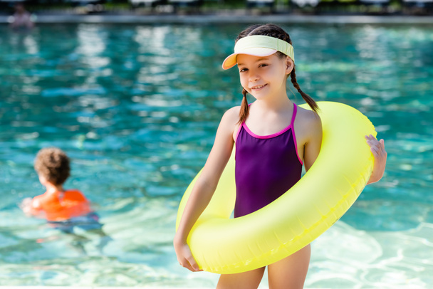 girl in swimsuit and sun visor cap posing with inflatable ring near pool - Photo, Image