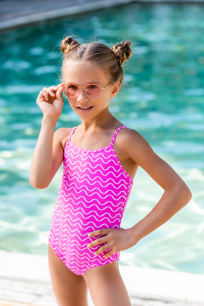 kind in badpak aanraken zonnebril terwijl poseren met de hand op heup in de buurt zwembad - Foto, afbeelding